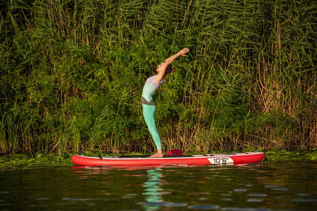 La giovane donna sta facendo yoga su un SUP stand up paddle su un bellissimo lago o fiume. Il concetto di uno stile di vita sano. Sport. Yoga. Passatempo