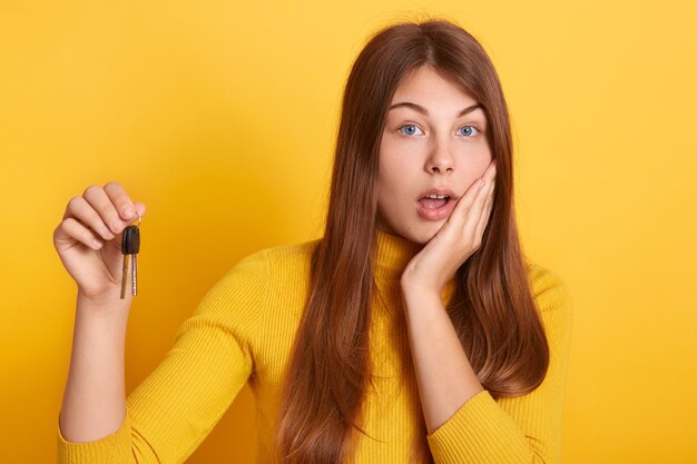 La giovane donna stupita che tiene in mano le chiavi di un'auto nuova o di un appartamento, ha un regalo inaspettato, posa isolata, indossa un abbigliamento casual