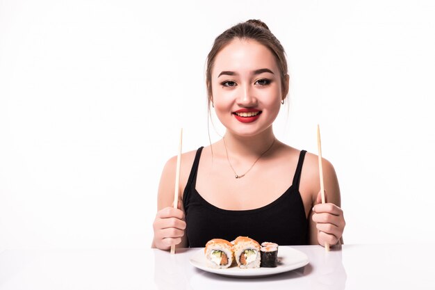 La giovane donna tenera è seduta al tavolo bianco e ha un piatto con sushi con bacchette di legno in entrambe le mani