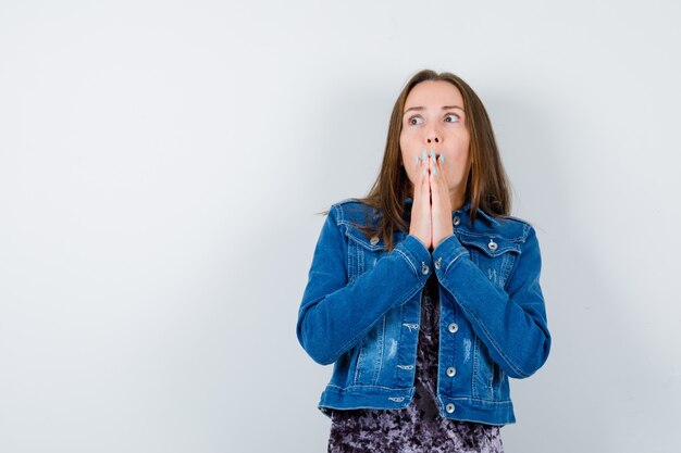 La giovane donna tiene i palmi premuti insieme in camicetta, giacca di jeans e sembra spaventata. vista frontale.