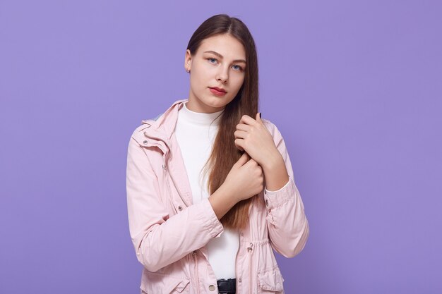 La giovane donna triste che si tocca i capelli mentre guarda direttamente, ha un'espressione facciale sconvolta, indossa una giacca rosa pallido e un dolcevita bianco, in piedi isolato sul muro lilla.