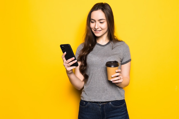 La giovane donna usa il telefono tenendo la tazza di caffè isolata sulla parete gialla