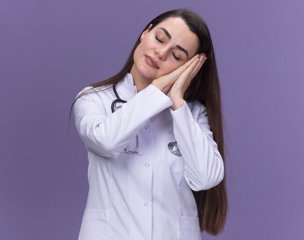 La giovane dottoressa assonnata che indossa un abito medico con lo stetoscopio mette la testa sulle mani isolate sulla parete viola con spazio per le copie