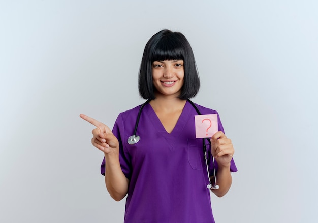 La giovane dottoressa bruna sorridente in uniforme con lo stetoscopio tiene la nota della domanda