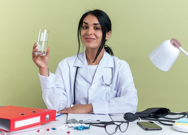 La giovane dottoressa contenta che indossa una veste medica con uno stetoscopio si siede alla scrivania con strumenti medici che tengono un bicchiere d'acqua isolato su una parete verde oliva