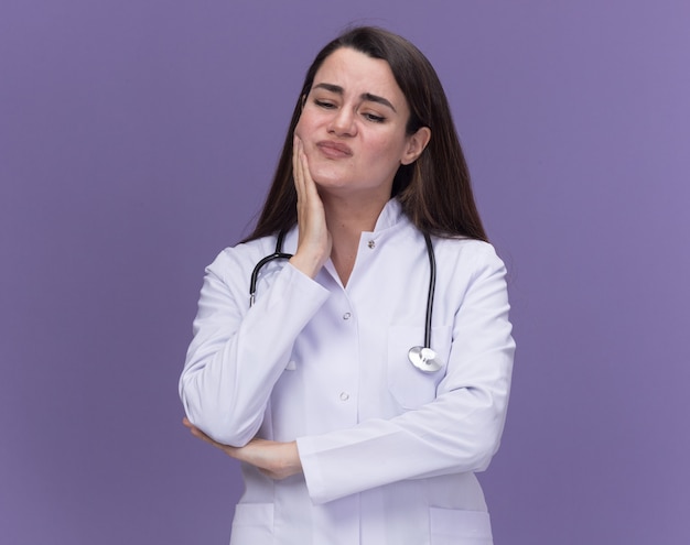 La giovane dottoressa scontenta che indossa l'abito medico con lo stetoscopio mette la mano sul mento e guarda giù isolata sulla parete viola con lo spazio della copia