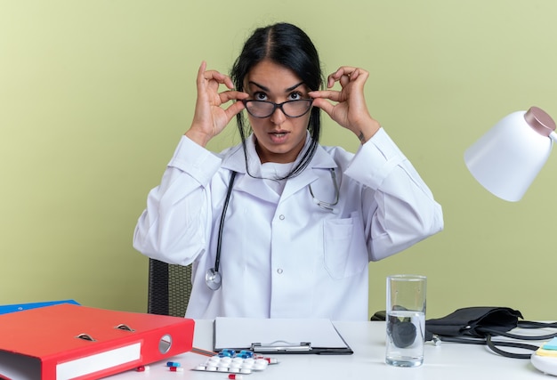 La giovane dottoressa sorpresa che indossa una veste medica con stetoscopio e occhiali si siede alla scrivania con strumenti medici isolati su sfondo verde oliva