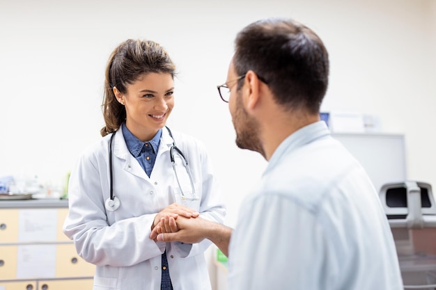 La giovane dottoressa tiene la mano dell'uomo caucasico paziente dà conforto espresso assistenza sanitaria simpatia aiuto medico fiducia supporto incoraggiare rassicurare il paziente sterile durante la visita medica