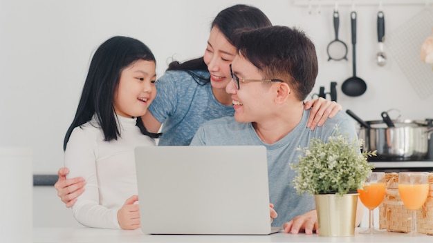 La giovane famiglia asiatica gode usando il computer portatile insieme a casa. Stile di vita giovane marito, moglie e figlia abbraccio felice e giocare dopo aver fatto colazione in cucina moderna a casa la mattina.
