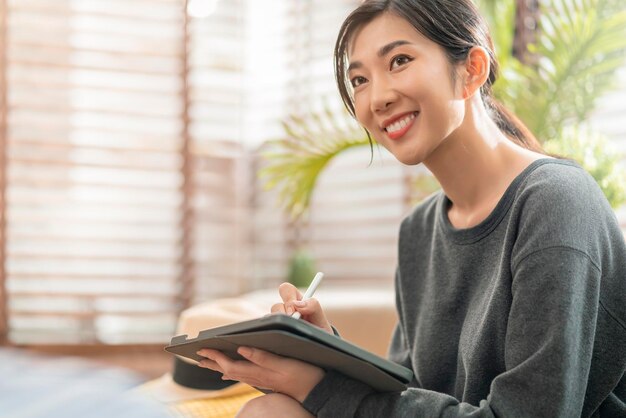 La giovane femmina asiatica attraente che lavora a casa usa la compressa che controlla il rapporto di pianificazione del grafico di dati di affari con il tempo libero si rilassa sul sofà nel concetto dell'isolato della casa del salone