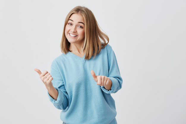 La giovane femmina caucasica felice emozionale con capelli biondi si è vestita in vestiti blu dando i suoi pollici in su, mostrando quanto è buono un prodotto. Ragazza graziosa che sorride pigramente con i denti. Gesti e linguaggio del corpo