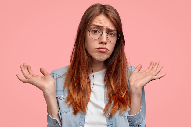 La giovane femmina di scontento perplesso con la pelle lentigginosa sta con i palmi aperti contro uno sfondo rosa, essendo confusa su qualcosa