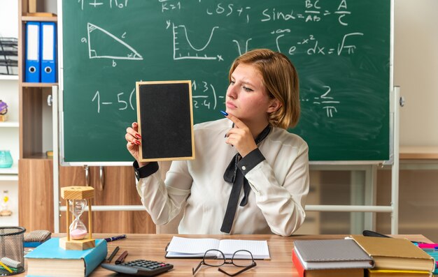 La giovane insegnante femminile si siede al tavolo con gli strumenti della scuola che tengono mini lavagna in classe