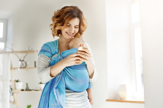 La giovane madre attraente sorride e guarda attraverso le foto del figlio sul telefono cellulare