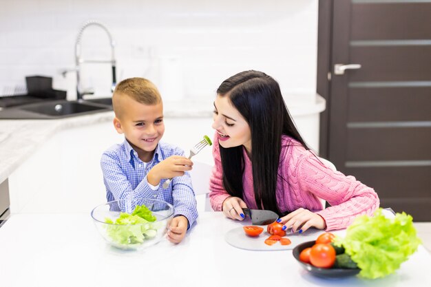 La giovane madre con il ragazzo del figlio che cucina la mamma dell'insalata ha affettato il figlio dell'alimento delle verdure alla madre che assaggia l'insalata. Famiglia felice cucinare cibo stile di vita godimento cucina