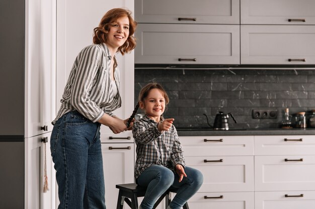 La giovane madre in camicia e jeans sta accanto a sua figlia che si siede sulla sedia.