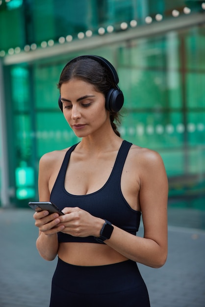 La giovane modella sportiva in cuffia cerca la canzone dalla playlist sfoglia la pagina web di fitness sullo smartphone legge la motivazione della vitalità indossa i resti della parte superiore tagliati dopo un duro allenamento si mantiene in salute