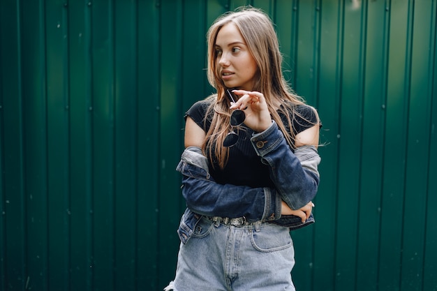 La giovane ragazza abbastanza attraente in vestiti del denim in vetri su una parete verde scuro semplice con il posto per testo.