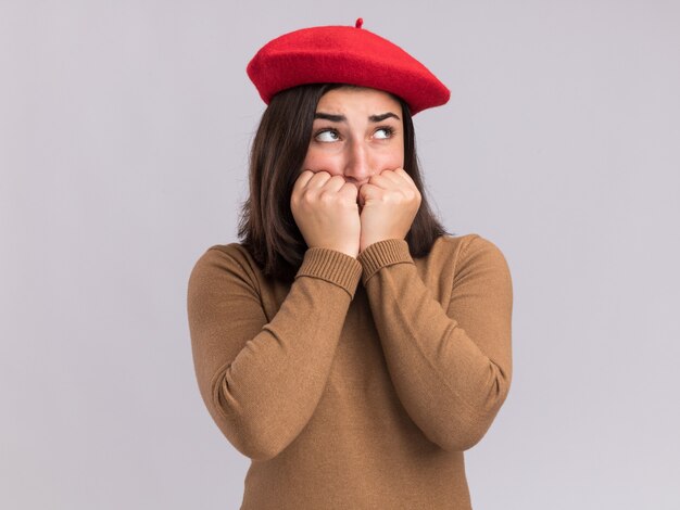 La giovane ragazza abbastanza caucasica ansiosa con il cappello del berretto mette i pugni sul mento che esamina il lato isolato sulla parete bianca con lo spazio della copia