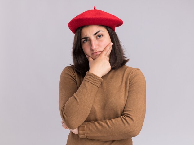 La giovane ragazza abbastanza caucasica confusa con il cappello del berretto tiene il mento isolato sulla parete bianca con lo spazio della copia