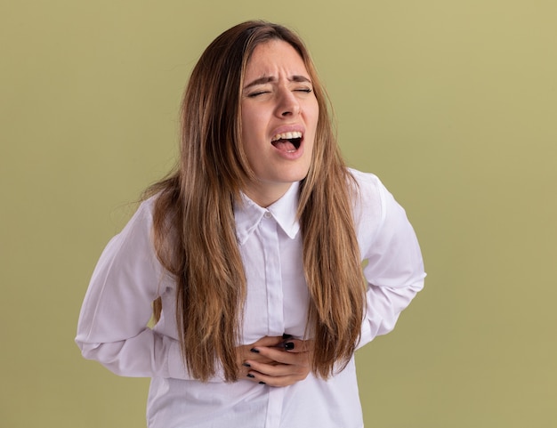 La giovane ragazza abbastanza caucasica dolorante mette le mani sulla pancia isolata sulla parete verde oliva con lo spazio della copia