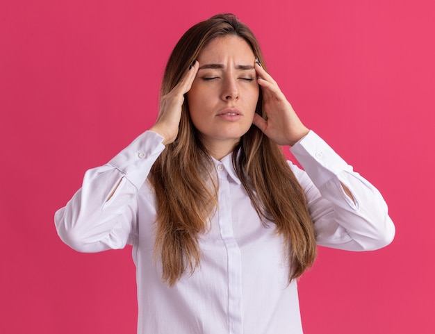 La giovane ragazza abbastanza caucasica dolorante mette le mani sulle tempie isolate sulla parete rosa con lo spazio della copia