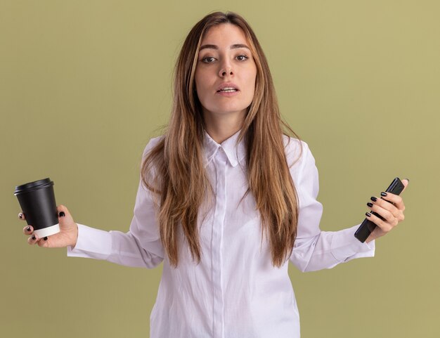 La giovane ragazza abbastanza caucasica sicura tiene il bicchiere di carta e il telefono