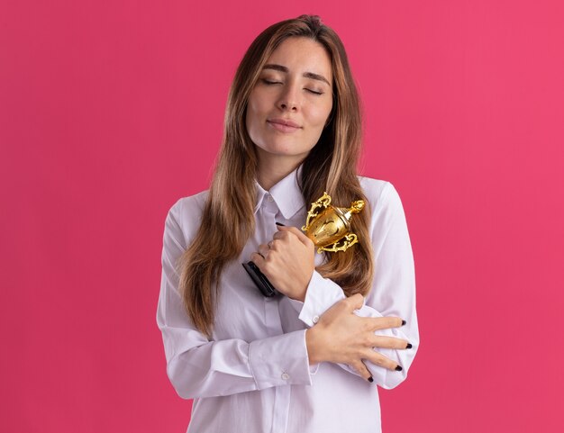 La giovane ragazza abbastanza caucasica soddisfatta sta con gli occhi chiusi che tengono la tazza del vincitore sul colore rosa