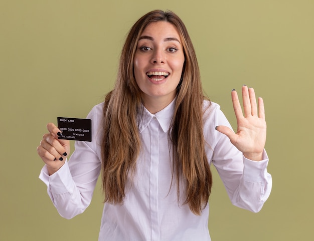 La giovane ragazza abbastanza caucasica soddisfatta tiene la carta di credito e gesti cinque su verde oliva