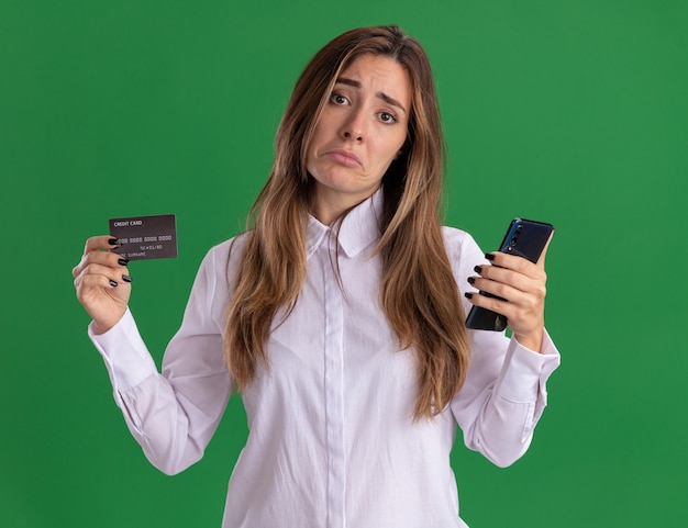 La giovane ragazza abbastanza caucasica triste tiene la carta di credito e il telefono isolati sulla parete verde con lo spazio della copia