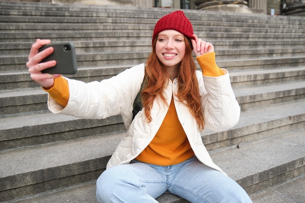 La giovane ragazza alla moda con il cappello rosso scatta foto sulla fotocamera dello smartphone e fa selfie mentre si siede sulle scale n