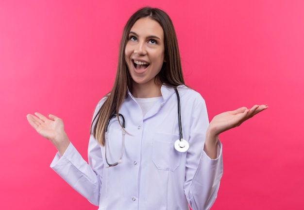 La giovane ragazza allegra del medico che porta l'abito medico dello stetoscopio diffonde le mani sulla parete rosa isolata