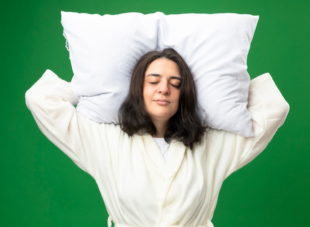 La giovane ragazza ammalata caucasica che indossa la veste che tiene il cuscino sotto la testa fa finta di dormire isolato su priorità bassa verde