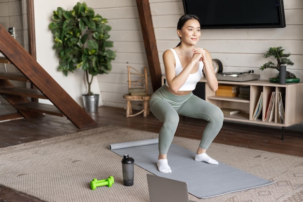 La giovane ragazza asiatica di forma fisica che fa l'allenamento di squat a casa segue l'istruttore di palestra di sport online in piedi su...