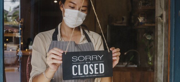 La giovane ragazza asiatica indossa una maschera facciale che trasforma un cartello da aperto a chiuso sul caffè con porta di vetro dopo la quarantena di blocco del coronavirus.