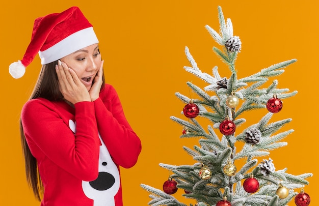 La giovane ragazza asiatica sorpresa che porta il cappello di natale con l'albero di Natale vicino in piedi maglione ha coperto le guance con le mani isolate su priorità bassa arancione