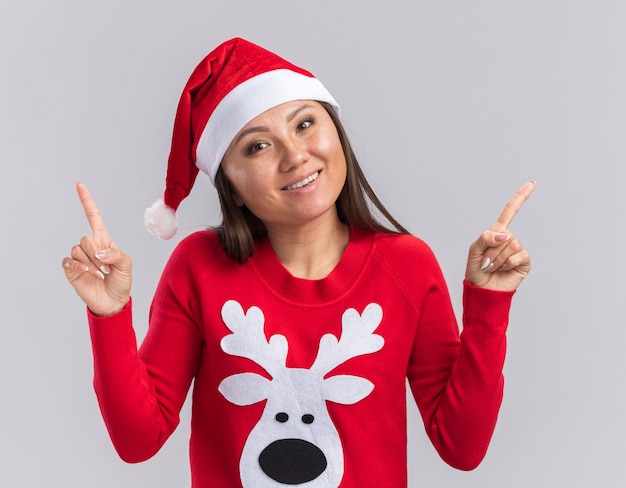 La giovane ragazza asiatica sorridente della testa di inclinazione che porta il cappello di natale con i punti del maglione in su isolato su fondo bianco