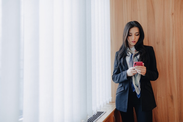 La giovane ragazza attraente emozionale in vestiti di stile di affari ad una finestra con un telefono in un ufficio o in una sala moderno