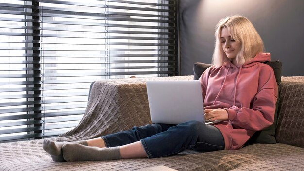 La giovane ragazza bionda del creatore di contenuti è sul suo laptop seduto sul divano vicino alla finestra