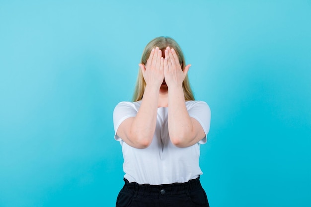 La giovane ragazza bionda si copre il viso con le mani su sfondo blu