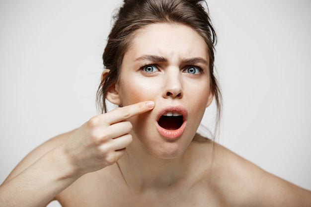 La giovane ragazza castana ha dispiaciuto della sua pelle del fronte dell'acne di problema sopra fondo bianco. Cosmetologia della salute e cura della pelle.