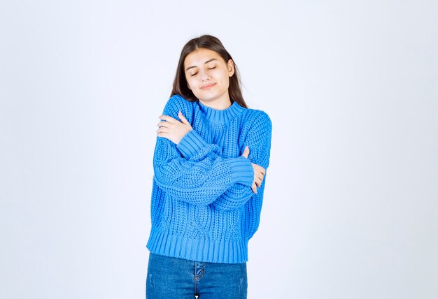 La giovane ragazza castana in maglione blu si abbraccia su bianco.