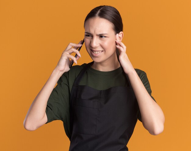 La giovane ragazza castana infastidita del barbiere in uniforme chiude l'orecchio e parla sul telefono che esamina il lato sull'arancio