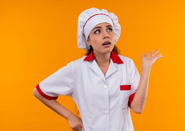 La giovane ragazza caucasica ansiosa del cuoco in uniforme del cuoco unico tiene la mano e cerca isolata su fondo arancio con lo spazio della copia