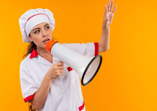 La giovane ragazza caucasica ansiosa del cuoco in uniforme dello chef tiene l'altoparlante e alza la mano isolata su fondo arancio con lo spazio della copia