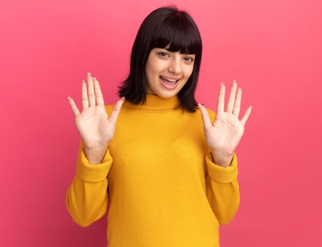 La giovane ragazza caucasica castana allegra sta con le mani alzate sul colore rosa