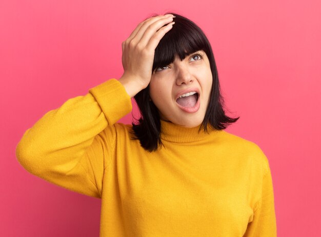 La giovane ragazza caucasica castana dispiaciuta mette la mano sulla testa e alza lo sguardo sul rosa