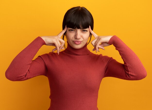 La giovane ragazza caucasica castana dispiaciuta mette le dita sulle tempie sull'arancia