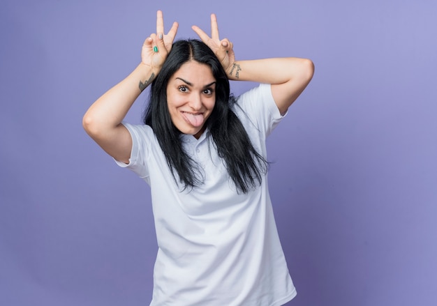 La giovane ragazza caucasica castana divertente tiene le mani sulla testa che gesturing le corna e sporge la lingua isolata sulla parete viola