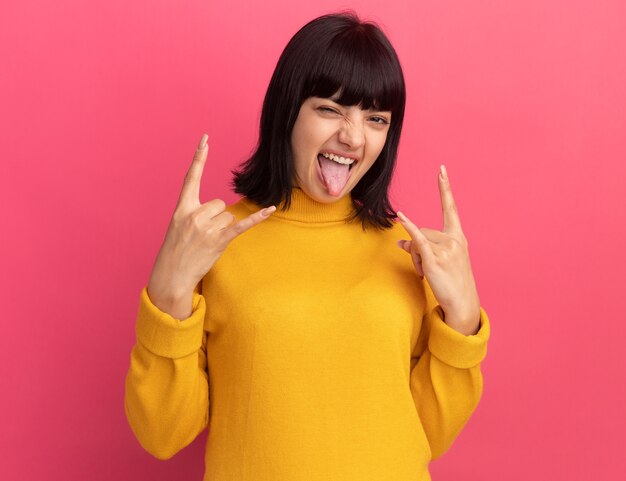La giovane ragazza caucasica castana gioiosa sporge la lingua e fa gesti con le corna con le mani isolate sulla parete rosa con spazio per le copie